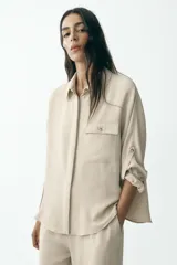 Camisa color marrón claro de cuello solapa y manga larga con trabilla y botón para remangar. Tiene un bolsillo de plastrón en el delantero, bajo asimétrico y cierre frontal con botones ocultos por solapa.