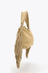 Mini bolso color beige de serraje, con flecos en los laterales, asa de mano trenzada y asa bandolera extraíble. Cierre con cremallera.