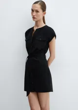 Vestido corto negro de tejido fluido, con cuello redondo y mangas cortas. Presenta trabillas, diseño evasé, dos bolsillos de solapa con botón en el pecho, lazo ajustable y cierre delantero con botones.