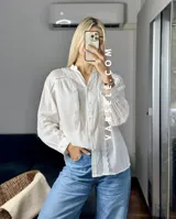 Camisa blanca de mangas largas con puños abullonados y detalles de encaje en cuello, mangas y ruedo.