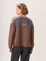 Buzo de manga larga con diseño de bloques de color y textura. El cuerpo principal es marrón, la espalda superior y hombros tienen un panel de tejido a rayas grises y azules, y los hombros superiores son de color beige rosado. Presenta costuras decorativas en rojo que separan los bloques de color.