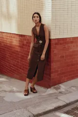 Vestido midi marrón oscuro, sin mangas, con cuello redondo y cierre frontal de botones decorativos. Presenta bolsillos de parche en la cadera y un cinturón elástico a tono con hebilla circular plateada y pedrería.