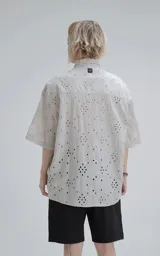 Camisa de manga corta color gris claro con cuello camisero y botones frontales. La tela presenta un bordado tipo broderie con motivos florales y geométricos.