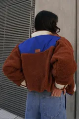 Campera marrón de tela tipo sherpa con cuello, puños y bolsillos de tela blanca. Tiene cierre con botones a presión y ribete azul.