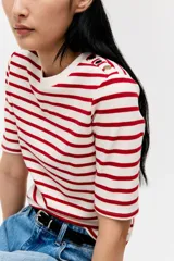 Remera entallada de punto a rayas rojas y blancas con cuello redondo y mangas cortas con botones dorados en los hombros.