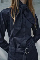 Camisa de denim azul oscuro con cuello con lazos, mangas largas con puños abotonados, bajo redondeado y cierre frontal con botones ocultos.