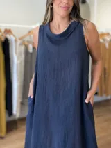 Vestido largo sin mangas color azul claro, confeccionado en lino, con cuello tipo buche o drapeado. Presenta un calce holgado y bolsillos laterales.