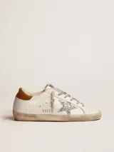 Championes Golden Goose modelo Super-Star confeccionados en cuero blanco con efecto desgastado. Destacan por su estrella lateral en glitter plateado, refuerzo del talón en cuero marrón y forro interior de lana tipo shearling. Incluyen cordones grises y suela de goma con acabado envejecido.