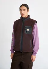 Chaleco de tejido polar tipo sherpa en color marrón oscuro, con cuello alto y cierre frontal completo. Presenta un bolsillo de parche en el pecho en tela técnica de contraste con etiqueta de la marca y bolsillos laterales con cremallera.