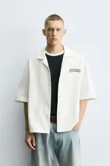 Camisa relaxed fit color blanco crudo, confeccionada en tejido de algodón con estructura. Cuello bowling y manga corta. Estampado frontal con la frase "ONLY DUST IS FOREVER" en negro. Cierre frontal de botonadura.