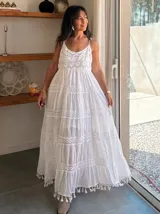 Vestido largo blanco de algodón con tirantes finos, escote en V con detalle de encaje calado y bordado. Falda con múltiples volados y terminaciones con flecos/borlas.