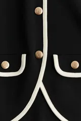 Saco negro de punto con ribetes blancos en cuello, bolsillos y borde inferior. Tiene corte entallado, cuello redondo, cierre frontal con botones dorados y solapas decorativas en el pecho y en la parte inferior.