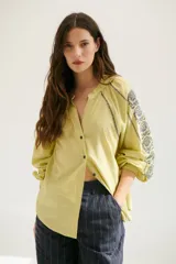 Camisa blanca de manga larga con bordados marrones en hombros y mangas, cuello en V y cierre frontal con botones.