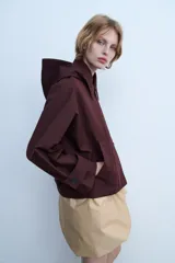 Trench corto color negro, con cuello solapa y capucha desmontable mediante botones. Manga larga tipo murciélago acabada en trabilla ajustable. Bolsillos laterales. Forro interior combinado. Cierre frontal con botones. Tejido repelente al agua.