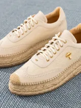 Zapatilla color crema con aplique de labial y logo dorado en el lateral, confeccionada en cuero y rafia, con suela de yute.