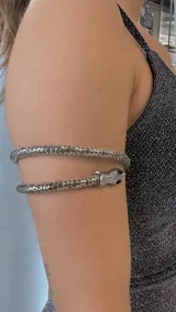 Brazalete rígido plateado con forma de serpiente enroscada en el brazo. La cabeza de la serpiente está decorada con strass y tiene ojos negros.