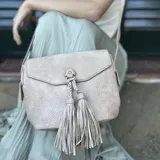 Bandolera color beige con tapa y broche magnético, apertura superior con cierre, detalle de trenzas artesanales y pompones en el frente, correa larga regulable y bolsillos internos.