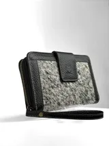 Billetera tipo clutch o cartera de mano, con cuerpo principal de cuero negro texturizado y panel central de pelo (efecto vaca) en tonos blanco y negro. Posee cierre de solapa con broche y correa de muñeca desmontable en cuero negro.