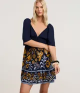 Vestido corto de viscosa con estampado floral y de follaje en tonos mostaza, blanco y azul marino. Presenta escote pronunciado en V con lazo y fruncido en el busto, mangas cortas abullonadas y elástico (lastex) en la espalda. La falda es evasé con un ruedo con volado.