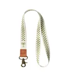 Llavero de cuello tipo lanyard con cinta de tela estampada con patrón de cuadros verdes y blancos. Presenta un detalle de parche en cuero sintético marrón con logo grabado, una estampa de palmera y un mosquetón metálico de color bronce.