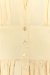 Vestido corto confeccionado en popelín de color amarillo pálido. Presenta un escote en pico pronunciado, diseño sin mangas y cierre frontal con botones. Incluye detalle de pliegues en la cintura que brindan volumen a la falda y bolsillos laterales ocultos en la costura.