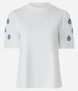 Remera blanca de algodón con cuello redondo, mangas cortas abullonadas y pequeños bordados florales azules en las mangas.