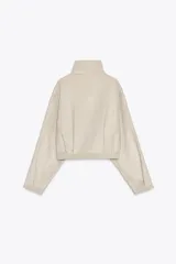 Cazadora corta color marrón claro, de cuello alto y manga larga. Tiene bolsillos laterales y cierre frontal con cremallera metálica.