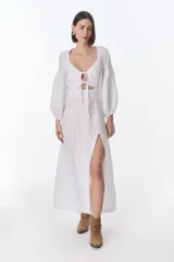 Vestido color beige de lino, con escote en V, mangas largas abullonadas y lazos frontales. Tiene corte a la cintura y falda larga con tajo frontal.