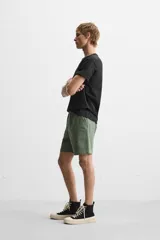Bermuda color verde militar de corte relajado, confeccionada en tejido de algodón. Tiene cintura elástica ajustable con cordón interior, bolsillos frontales y bolsillo trasero de parche.