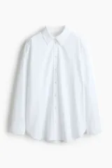 Camisa blanca de algodón con cuello clásico, abotonada adelante y corte holgado. Tiene mangas largas con puños sencillos y bajo redondeado con espalda algo más larga.