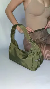 Cartera negra de lona semi impermeable con cierre metálico, forrería de gross y bolsillo interno. Tiene tira regulable y etiqueta blanca con texto en el frente.