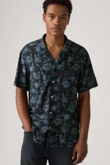 Camisa de hombre de manga corta con estampado floral en tonos azules sobre fondo negro. Tiene cuello tipo cubano, cierre frontal con botones y un pequeño logo rojo bordado en el pecho.