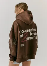 Buzo marrón chocolate de frisa con capucha, bolsillos delanteros y corte oversize. Estampa en frente y espalda con la frase "co-creator of love & dreams" y el logo de la marca Renée House.