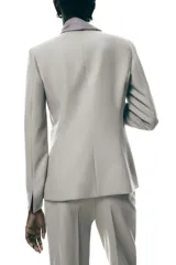 Blazer color beige con cuello solapa, manga larga con abertura y detalle de lazo gris en el frente.