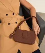 Bolso baguette pequeño color marrón con textura en relieve, correa ajustable, cierre por cremallera y solapa con detalle de colgante en el lateral.