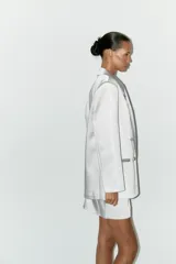Blazer oversize de satén color blanco, con cuello solapa, escote pico cruzado, manga larga con hombreras, bolsillos de doble vivo y abertura en la espalda. Cierre frontal cruzado con botones forrados.