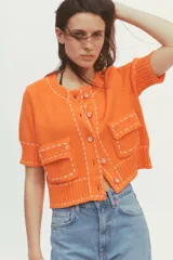 Chaqueta naranja de punto con cuello redondo y manga corta. Presenta bolsillos delanteros de parche con solapa y cierre frontal con botones. Detalle de pespuntes blancos a contraste.