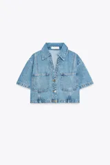 Camisa de denim celeste de corte crop, con cuello solapa, escote en pico, manga corta y dos bolsillos delanteros con botón. Cierre frontal con botones metálicos.