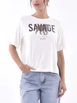Remera blanca de manga corta con cuello redondo y estampado frontal que dice "SAVAGE" con una figura de animal (posiblemente un leopardo o jaguar) hecho de lentejuelas o material brillante. Debajo del estampado principal se lee "LIFE GOES ON ENJOY".