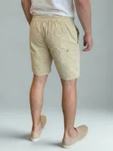 Bermuda color beige claro, de corte casual, con cintura elástica y cordón ajustable. Presenta un bolsillo trasero con ribete y un pequeño detalle de etiqueta marrón en el bolsillo trasero.