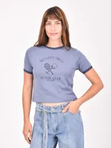 Remera ajustada color azul grisáceo, con cuello redondo y ribete en contraste en el cuello y las mangas. Presenta un estampado frontal con texto que dice "Beverly Hills Tennis Club Est. 1985" y un gráfico de una raqueta de tenis y una copa de vino cruzadas.