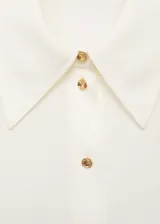 Camisa blanca de tejido fluido, con cuello camisero, cierre frontal con botones y mangas largas con puños abotonados.