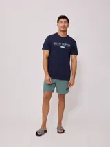 Remera color crema de manga corta con estampado frontal con el texto "Rusty Supply Surfboards" en color rojo y negro.