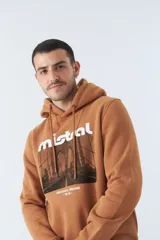 Buzo marrón con capucha y bolsillo canguro. Presenta un estampado frontal con el logo de la marca Mistral y una imagen de un arco arquitectónico. Confeccionado en algodón y poliéster.