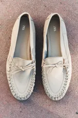 Zapato plano tipo mocasín color crema, confeccionado en cuero, con detalle de lazo con pasador y fruncido en la parte delantera. Pespunte en el corte y acabado en punta redonda.
