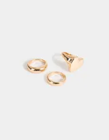 Set de tres anillos dorados de metal con diferentes diseños: uno liso, uno con forma de corazón y uno con forma de domo.