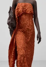 Vestido midi color ladrillo con textura arrugada, de tirantes finos y escote recto con volante. Presenta un corte asimétrico con una pieza larga que cae lateralmente.