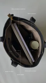 Cartera tipo tote de cuero graneado color negro, con textura uniforme y suave al tacto. Tiene manijas de 55 cm, bolsillo frontal con broche imantado, bolsillo trasero con cierre metálico, cierre principal metálico, forro interior y tres bolsillos internos (uno con cierre, uno plaqué y uno tipo sobre con solapa y botón).