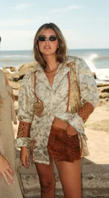 Blazer de corte recto confeccionado en brocado con diseño floral y paneles laterales de lentejuelas doradas. Presenta mangas con volumen en la parte superior, detalles de calados geométricos en los antebrazos y puños con lazos. Incluye recortes decorativos en contraste y cierre frontal con botón.