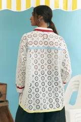 Camisa blanca oversize de algodón con bordado calado tipo broderie, cuello camisero con ribete rojo, cierre frontal con botones y mangas largas con puños con ribete rojo. Presenta una franja vertical con ribete celeste y otra con ribete amarillo.
